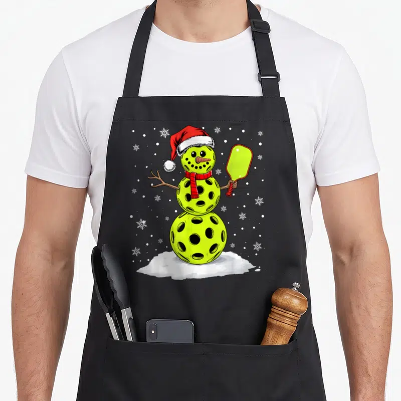 Hat snowman apron - Lime green pickleball snowman wearing red Santa hat and scarf holding a paddle on white snow — shown on a black cotton canvas apron against a white background