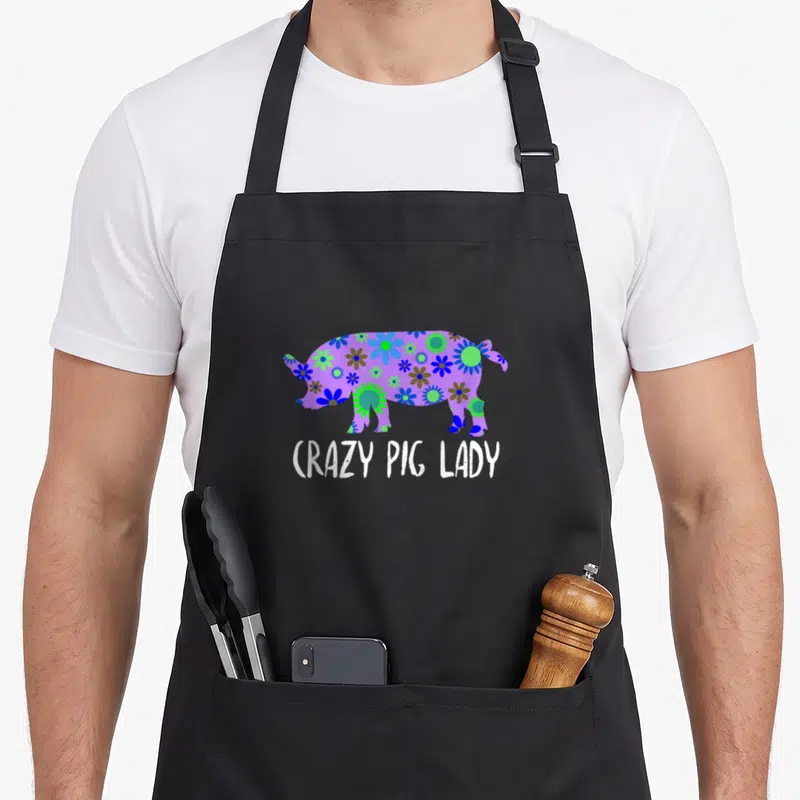 Farming floral apron - Purple pig silhouette filled with blue and green flowers above white text reading CRAZY PIG LADY — shown on a black cotton canvas apron against a white background