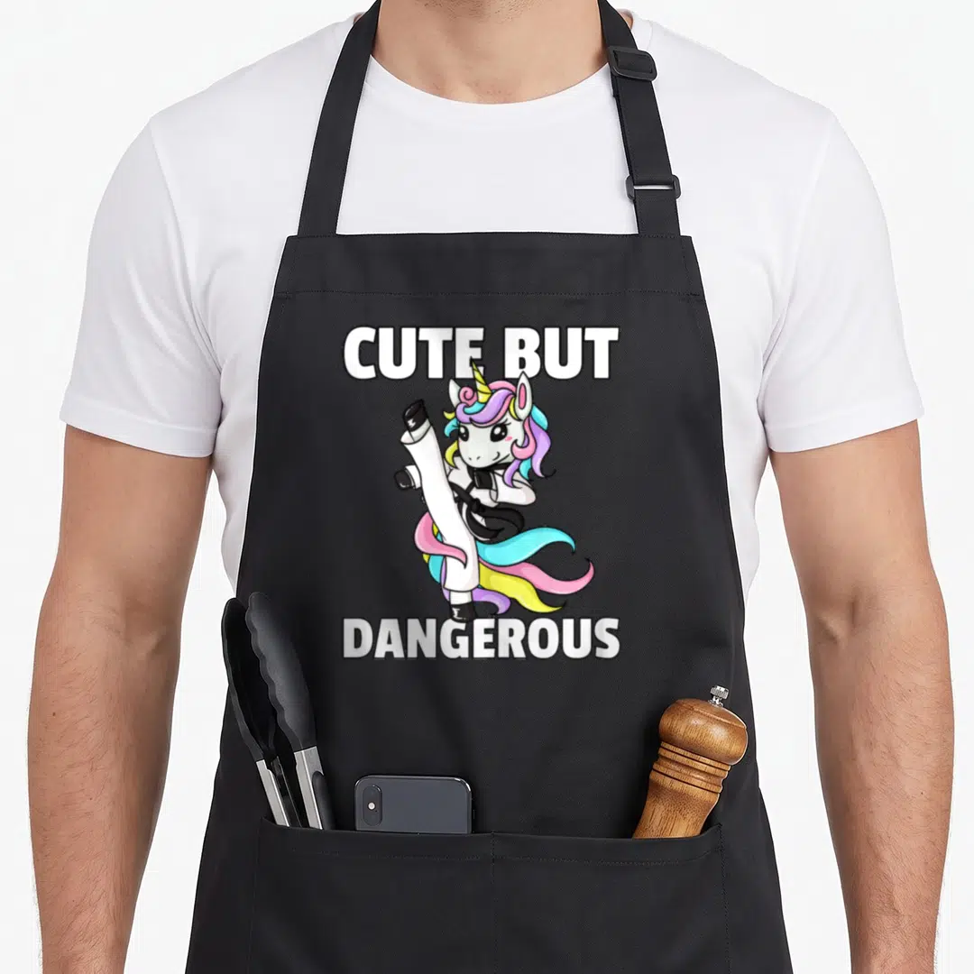Taekwondo unicorn apron - Cute But Dangerous text above cartoon unicorn kicking in white gi with rainbow mane — shown on a black cotton canvas apron against a white background