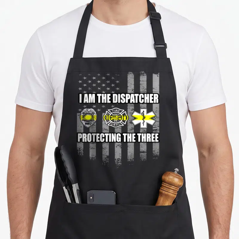 Dispatcher tree apron - I AM THE DISPATCHER text above police fire and EMS badges on distressed flag background — shown on a black cotton canvas apron against a white background