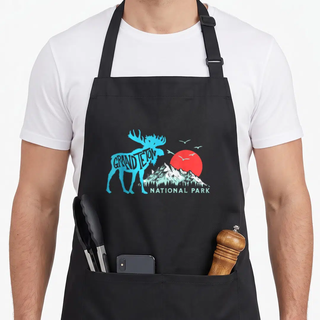 Moose grand teton national park apron - Blue moose silhouette with Grand Teton text above red sun and mountains — shown on a black cotton canvas apron against a white background