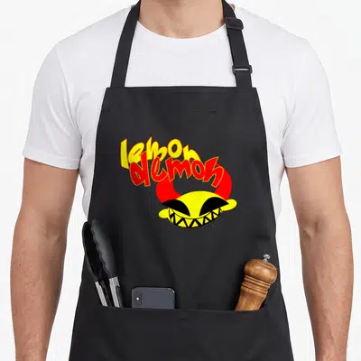 Demon lemon apron - Stacked lemon demon text over yellow cartoon head with red horns and black grin — shown on a black cotton canvas apron against a white background