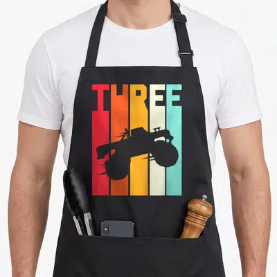 3rd truck aprons - Grey monster truck silhouette over red orange yellow cream and teal vertical stripes spelling THREE — shown on a black cotton canvas apron against a white background
