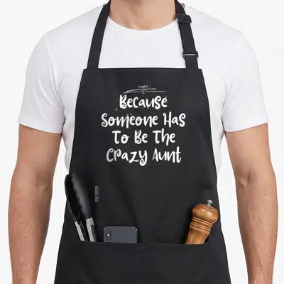 Someone christmas apron - White stacked text Because Someone Has To Be The Crazy Aunt with small heart above — shown on a black cotton canvas apron against a white background