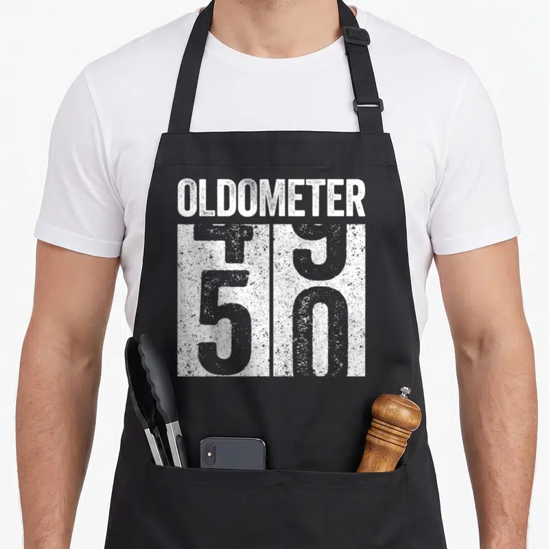 Oldometer birthday apron - Distressed white OLDOMETER text above grey 50 odometer graphic on black fabric — shown on a black cotton canvas apron against a white background