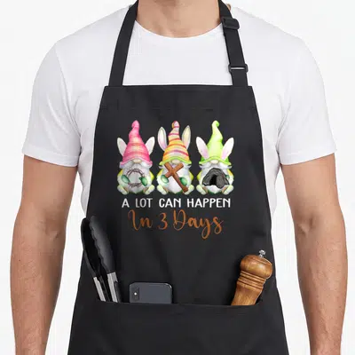 Gnome easter apron - Three watercolor gnomes with bunny ears holding thorns, cross, and stone with text A LOT CAN — shown on a black cotton canvas apron against a white background