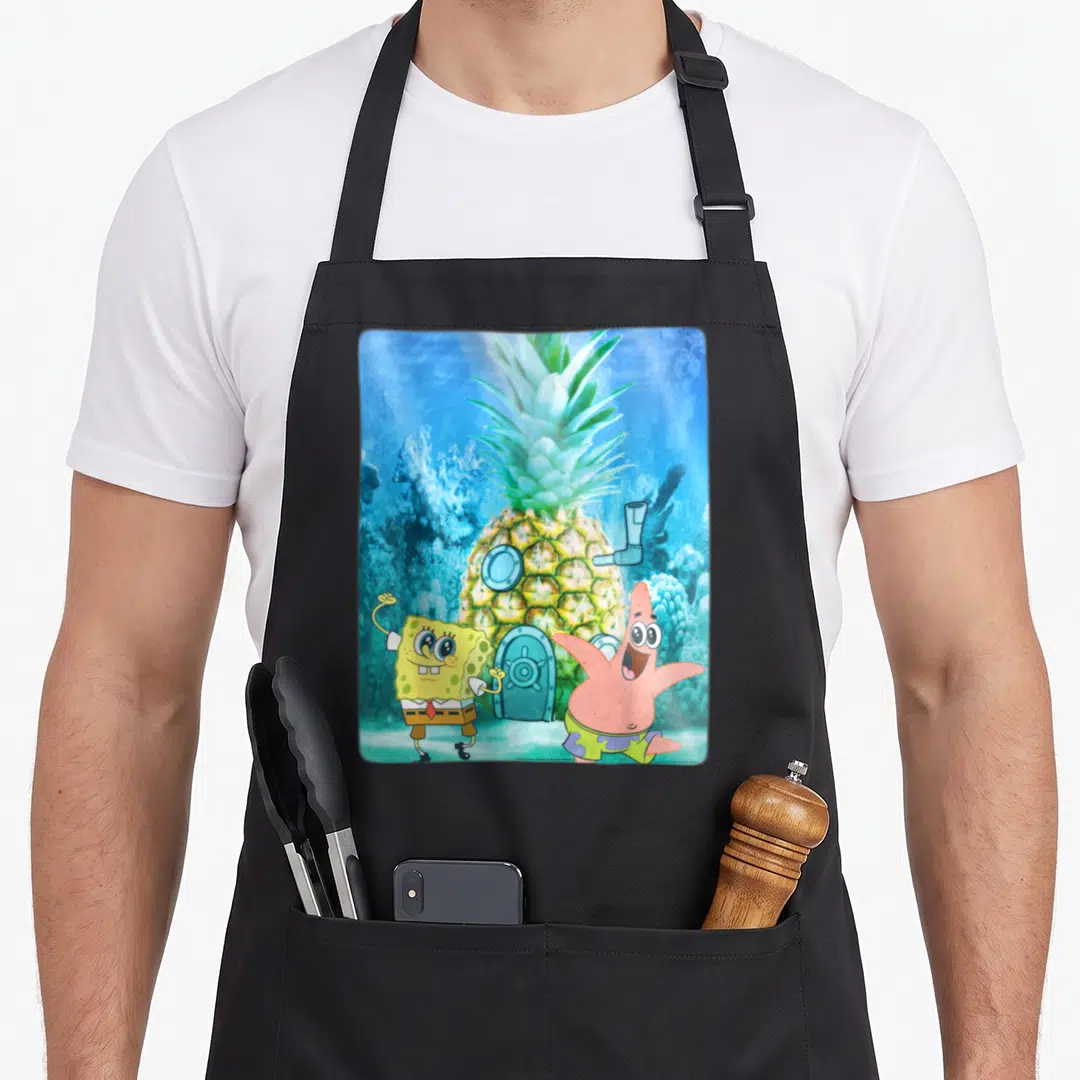 Squarepant fish apron - Yellow sponge and pink starfish standing in front of a pineapple house with blue underwater — shown on a black cotton canvas apron against a white background