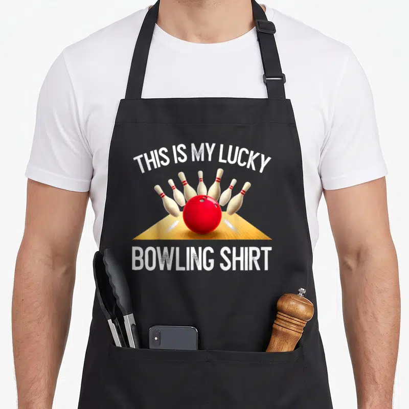 Lucky retro bowling apron - Red bowling ball hitting pins with THIS IS MY LUCKY BOWLING SHIRT text — shown on a black cotton canvas apron against a white background