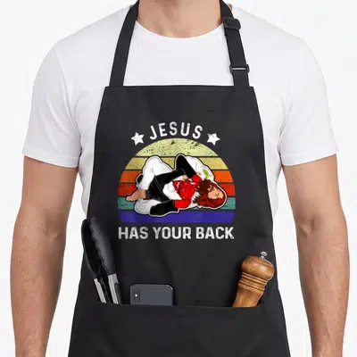 Jiu brazilian apron - Cartoon Jesus in gi pinning red devil under arched JESUS HAS YOUR BACK text on retro sunset — shown on a black cotton canvas apron against a white background