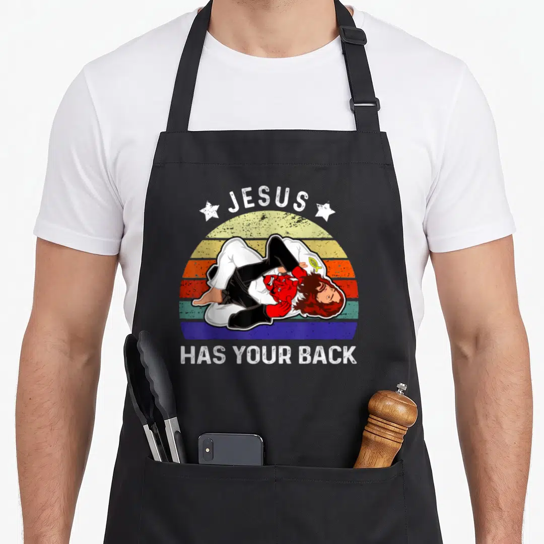 Jiu brazilian apron - Cartoon Jesus in gi pinning red devil under arched JESUS HAS YOUR BACK text on retro sunset — shown on a black cotton canvas apron against a white background