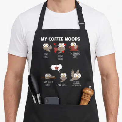 Coffee owl apron - Six cartoon owls with captions about coffee moods including love, drinking, and missing coffee — shown on a black cotton canvas apron against a white background