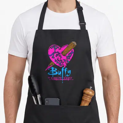 Buffy colorful aprons - Pink heart pierced by brown stake with blue veins and stacked cartoon text below — shown on a black cotton canvas apron against a white background