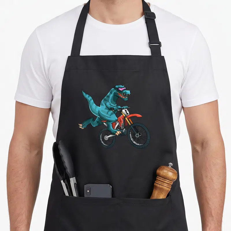 Dino bike apron - Blue dinosaur in purple goggles riding red and yellow dirt bike with number 1 — shown on a black cotton canvas apron against a white background