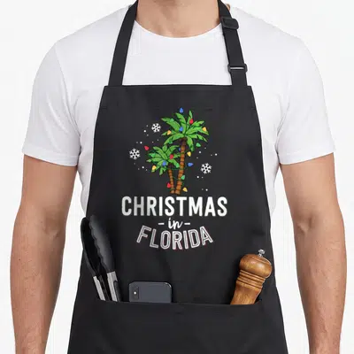 Florida christmas tree apron - Two palm trees wrapped in multicolored lights above stacked text reading CHRISTMAS — shown on a black cotton canvas apron against a white background