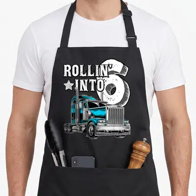 6th semi truck apron - Blue semi-truck with ROLLIN INTO text and tire shaped number 6 — shown on a black cotton canvas apron against a white background
