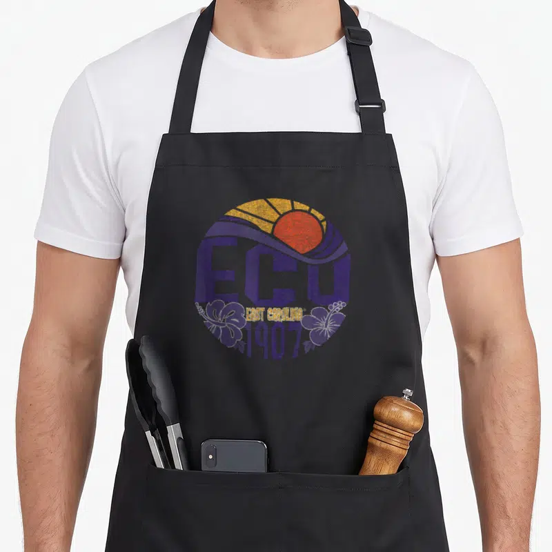 Shine carolina girl apron - Retro ECU circle with sun, waves, hibiscus, and 1907 text in purple orange yellow — shown on a black cotton canvas apron against a white background