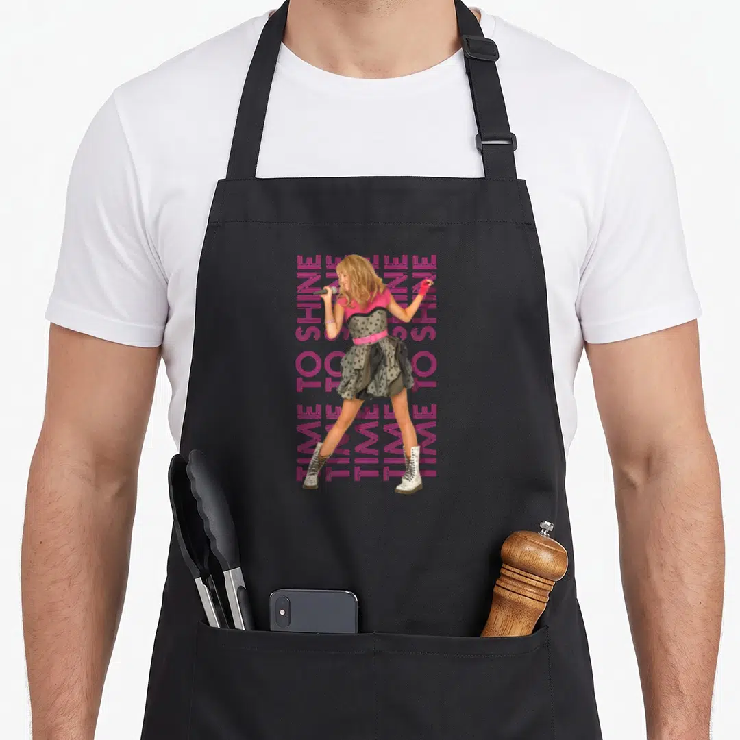 Shine montana girl apron - Woman standing with pink TIME TO SHINE text repeated four times vertically in grey and white — shown on a black cotton canvas apron against a white background