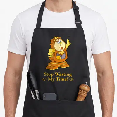 Cogsworth novelty apron - Anthropomorphic brown clock with monocle and yellow text reading Stop Wasting My Time — shown on a black cotton canvas apron against a white background