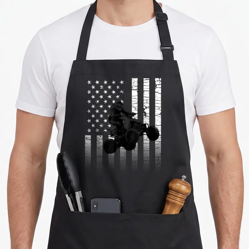 Atv american flag apron - Distressed black and white ATV rider jumping in front of US flag silhouette — shown on a black cotton canvas apron against a white background