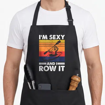 Row kayak building apron - Retro kayaker silhouette on yellow orange red blue stripes with white I'm sexy row it text — shown on a black cotton canvas apron against a white background