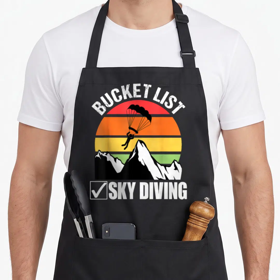 Sky bucket apron - Retro skydiver silhouette over striped sun and mountains with BUCKET LIST SKY DIVING text — shown on a black cotton canvas apron against a white background