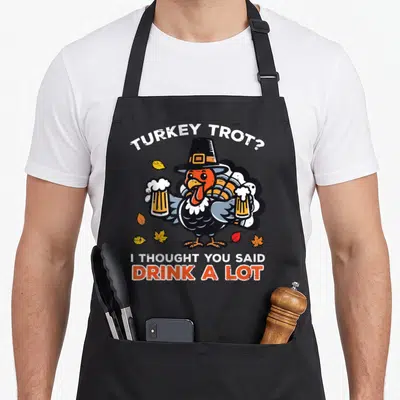 Thought thanksgiving apron - Cartoon turkey in pilgrim hat holding beer glasses with TURKEY TROT pun text — shown on a black cotton canvas apron against a white background