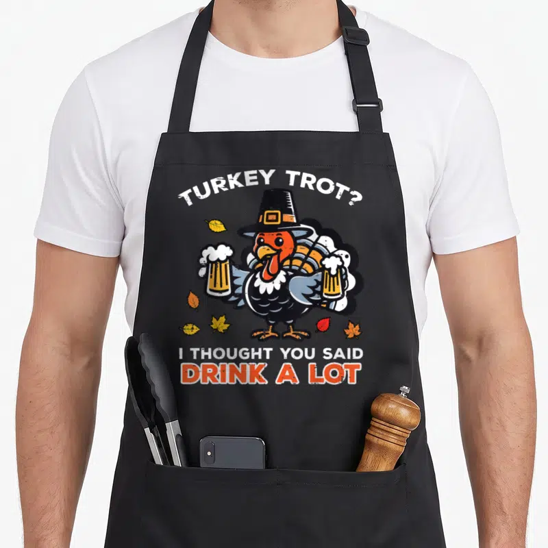 Thought thanksgiving apron - Cartoon turkey in pilgrim hat holding beer glasses with TURKEY TROT pun text — shown on a black cotton canvas apron against a white background