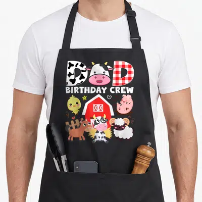 Theme farm apron - DAD BIRTHDAY CREW text with cow head and cartoon pig, horse, sheep, chick, and barn — shown on a black cotton canvas apron against a white background