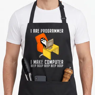 Programmer programming nerd apron - Penguin in sunglasses at yellow desk with I ARE PROGRAMMER text — shown on a black cotton canvas apron against a white background