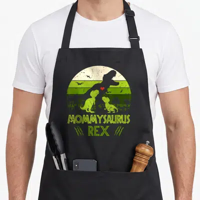 Mommysaurus dinosaur apron - Retro T-Rex family silhouette in green stripes with MOMMYSAURUS REX text and claw marks — shown on a black cotton canvas apron against a white background