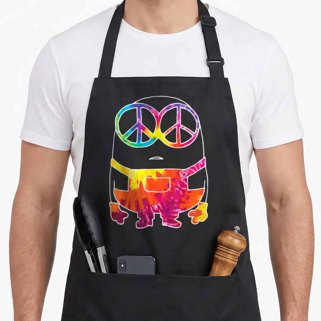 Despicable tie dye apron - Grey Minion outline with tie-dye peace sign goggles and colorful overalls — shown on a black cotton canvas apron against a white background