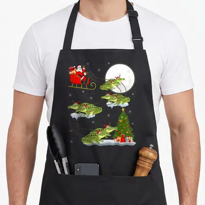 Lighting christmas tree apron - Santa in red sleigh pulled by eight green crocodiles with antlers over moonlit sky — shown on a black cotton canvas apron against a white background