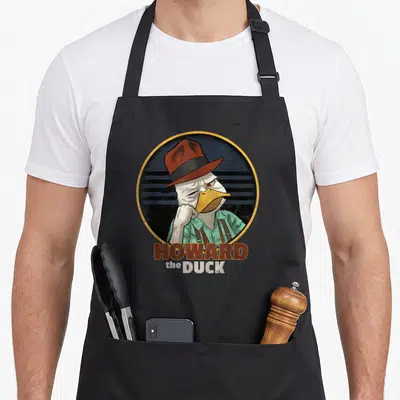 Howard duck apron - Cartoon duck in brown hat and green shirt inside yellow-bordered dark blue circle with text below — shown on a black cotton canvas apron against a white background