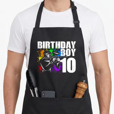 Paintball 10th birthday apron - Cartoon paintball player with white BIRTHDAY BOY 10 text and colorful splatters — shown on a black cotton canvas apron against a white background