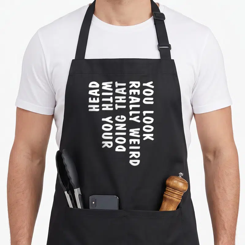Doing weird aprons - Vertical white text reading You Look Really Weird Doing That With Your Head on dark background — shown on a black cotton canvas apron against a white background