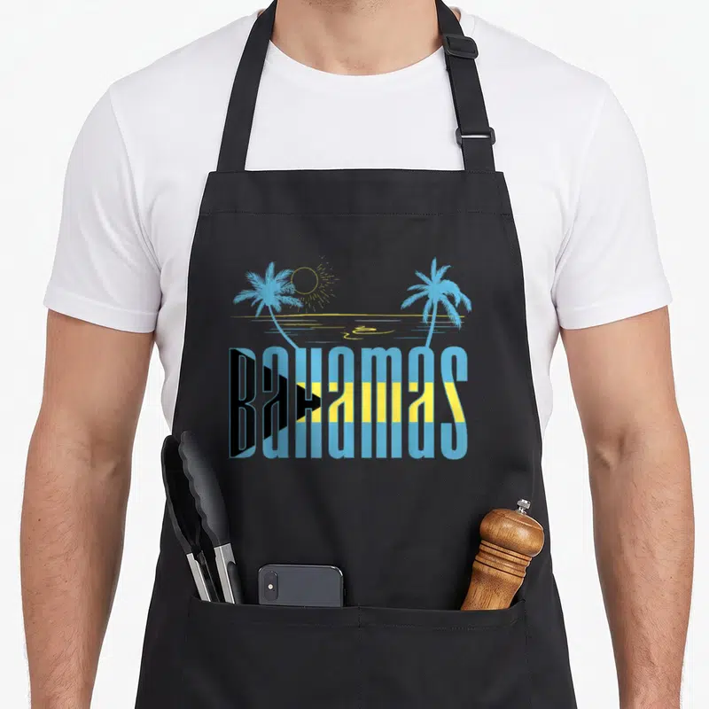 Nassau bahamian apron - BAHAMAS text in flag colors with two palm trees and a sun above an ocean line — shown on a black cotton canvas apron against a white background