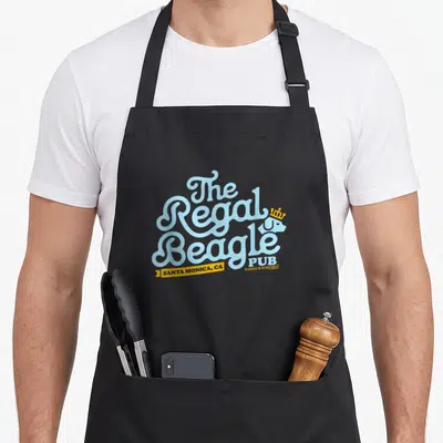 Regal beagle apron - Stacked light blue text The Regal Beagle PUB with white crowned beagle and yellow Santa Monica — shown on a black cotton canvas apron against a white background