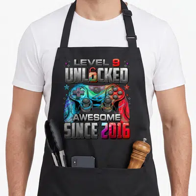 9th birthday gaming aprons - Colorful game controller with Level 9 Unlocked text and 2016 date — shown on a black cotton canvas apron against a white background