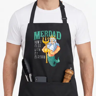 Merdad birthday apron - Cartoon merman holding gold trident with arched teal MERDAD text and stacked white slogan — shown on a black cotton canvas apron against a white background