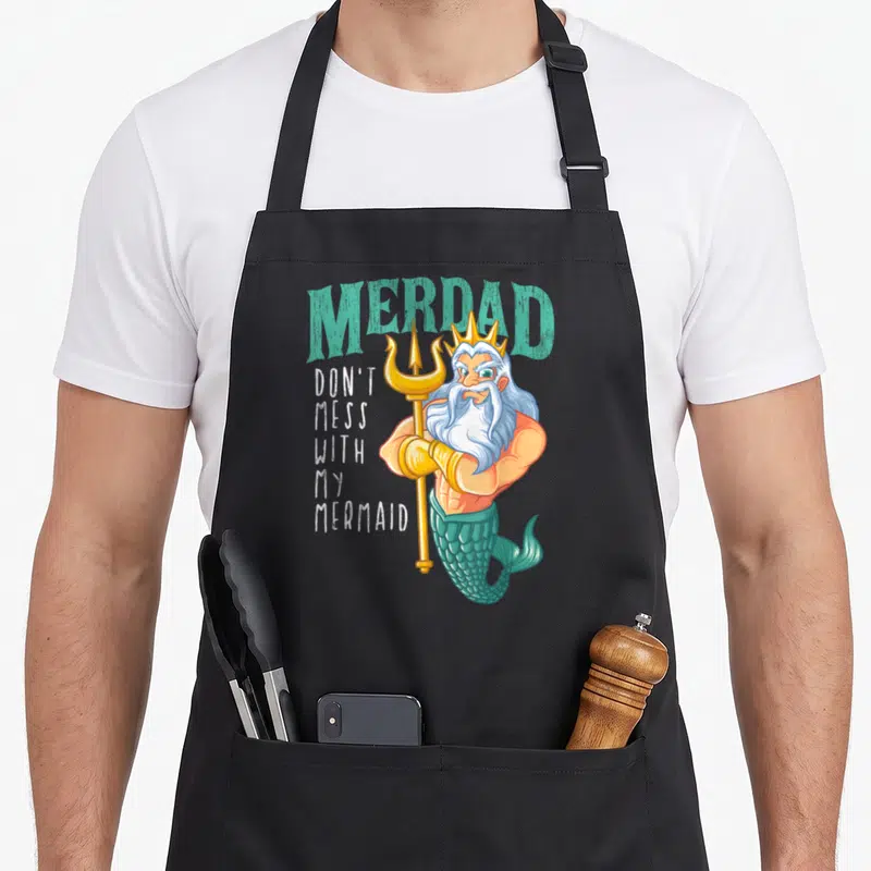 Merdad birthday apron - Cartoon merman holding gold trident with arched teal MERDAD text and stacked white slogan — shown on a black cotton canvas apron against a white background