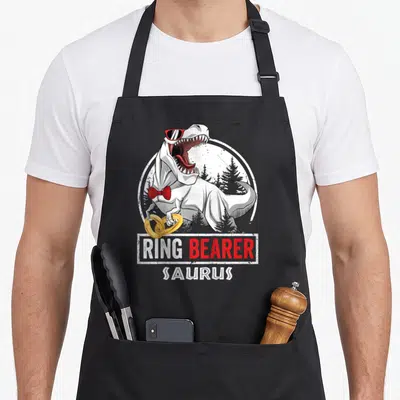 Ring wedding apron - T-Rex in sunglasses and red bow tie holding two gold rings below RING BEARER SAURUS text — shown on a black cotton canvas apron against a white background