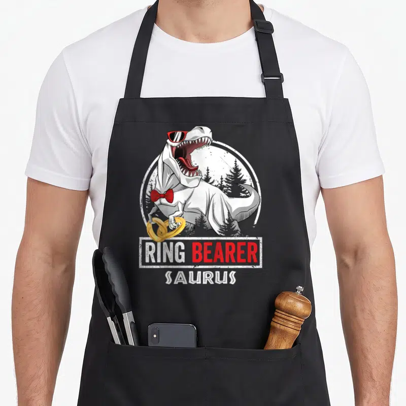 Ring wedding apron - T-Rex in sunglasses and red bow tie holding two gold rings below RING BEARER SAURUS text — shown on a black cotton canvas apron against a white background