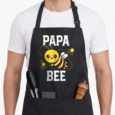 Outfit daddy apron - Cartoon bee between PAPA and BEE text with yellow honeycombs — shown on a black cotton canvas apron against a white background