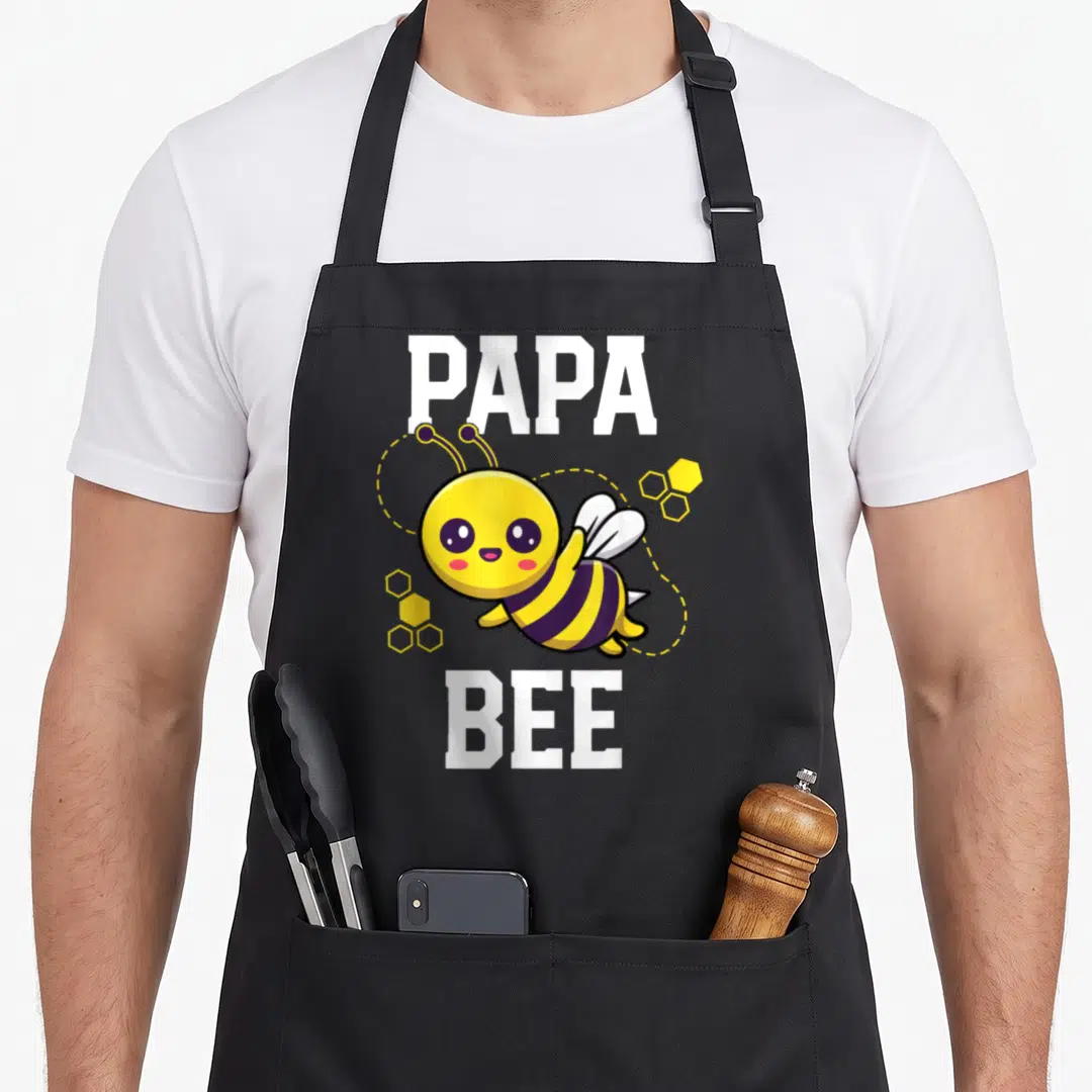 Outfit daddy apron - Cartoon bee between PAPA and BEE text with yellow honeycombs — shown on a black cotton canvas apron against a white background