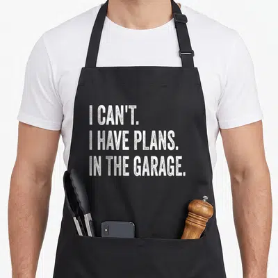 Plan garage apron - White stacked text reading I CANT I HAVE PLANS IN THE GARAGE on black fabric — shown on a black cotton canvas apron against a white background
