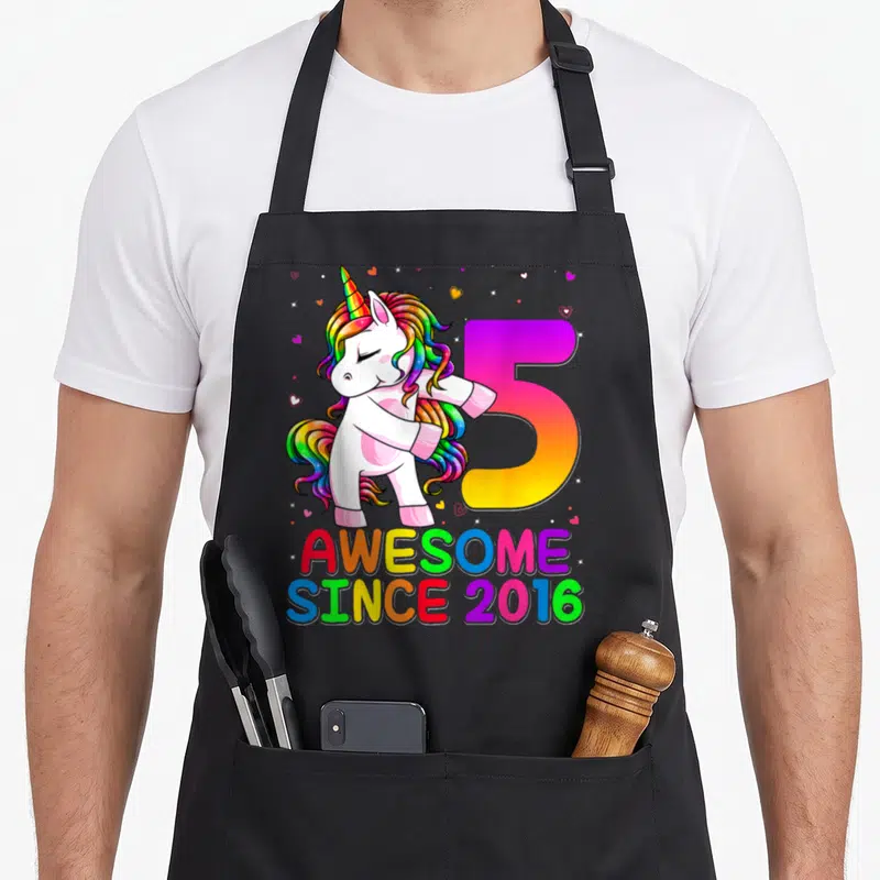 Flossing unicorn apron - Cartoon unicorn dancing next to rainbow number 5 and text AWESOME SINCE 2016 — shown on a black cotton canvas apron against a white background
