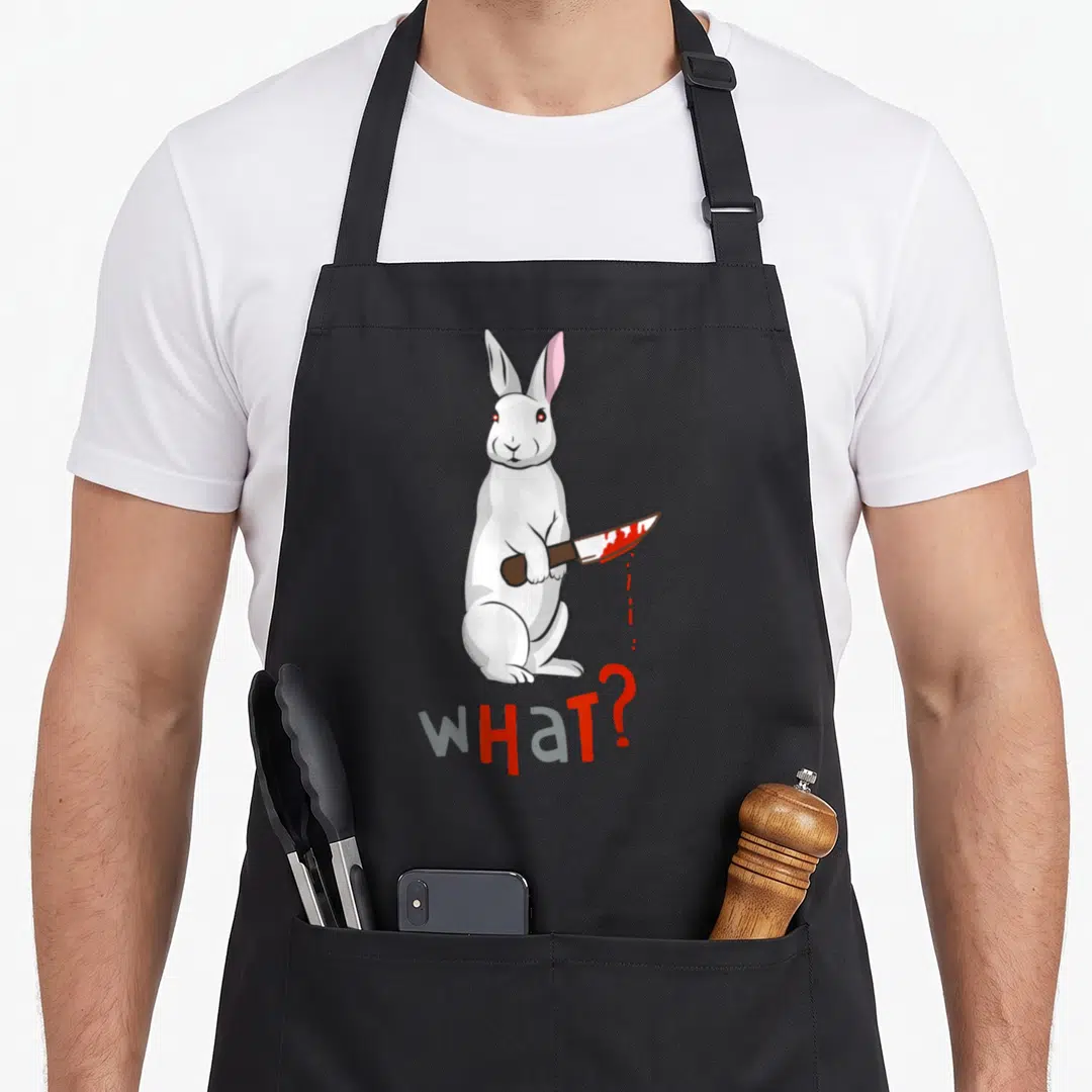 Scary rabbit apron - White cartoon rabbit holding bloody knife above WHAT? text in white grey red — shown on a black cotton canvas apron against a white background