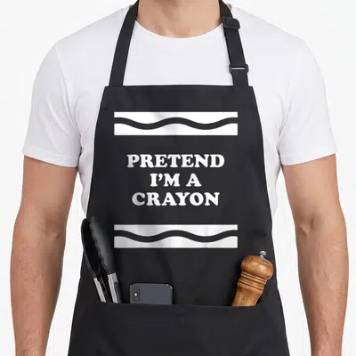 Pretend crayon halloween apron - White text PRETEND I'M A CRAYON between two wavy horizontal bands — shown on a black cotton canvas apron against a white background