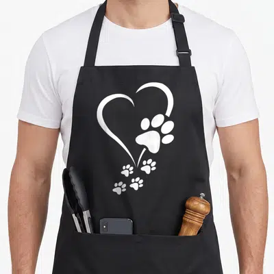 Baby paw print apron - White heart outline with one large paw print and four smaller prints trailing down on black — shown on a black cotton canvas apron against a white background