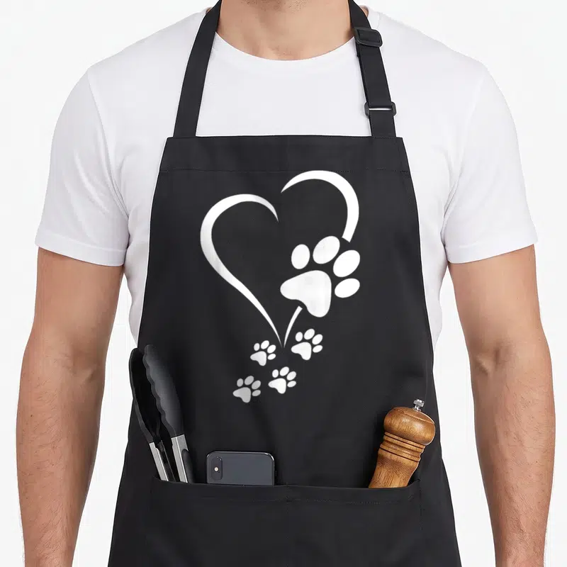 Baby paw print apron - White heart outline with one large paw print and four smaller prints trailing down on black — shown on a black cotton canvas apron against a white background